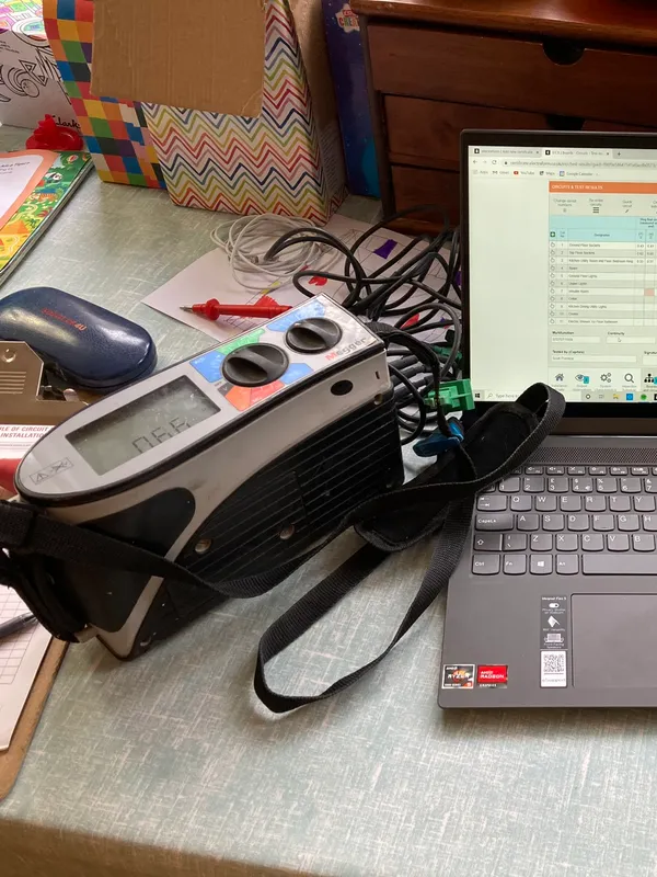 Laptop running electrical certification software beside a Megger multifunction tester during an EICR